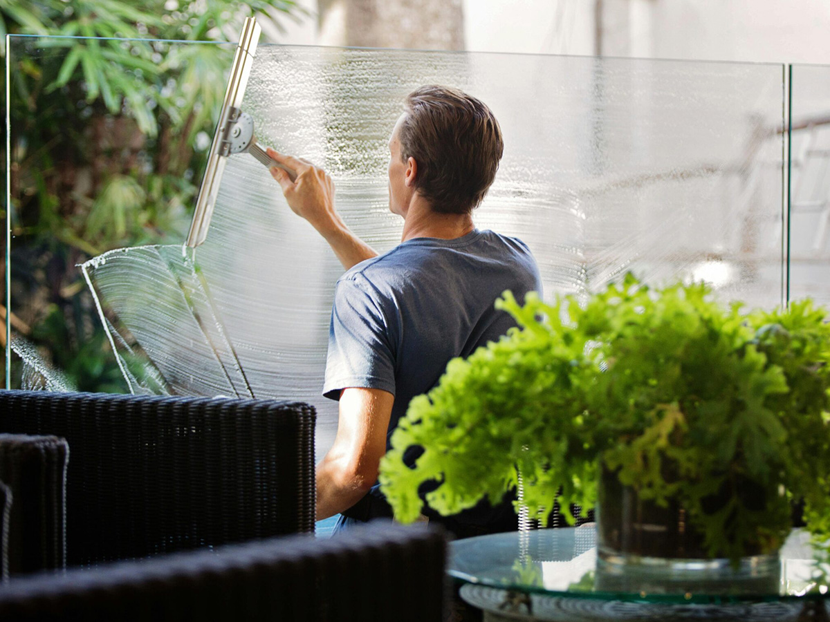 Mann in blauem T-Shirt putzt ein Glasgeländer auf einer sonnigen Terrasse mit Abzieher und Sprühflasche. Im Hintergrund sind Gartenmöbel.